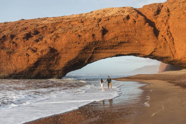 Couple in love walking under huge arch on Legzira ocean beach at sunset. Famous and popular landmark in Morocco.
